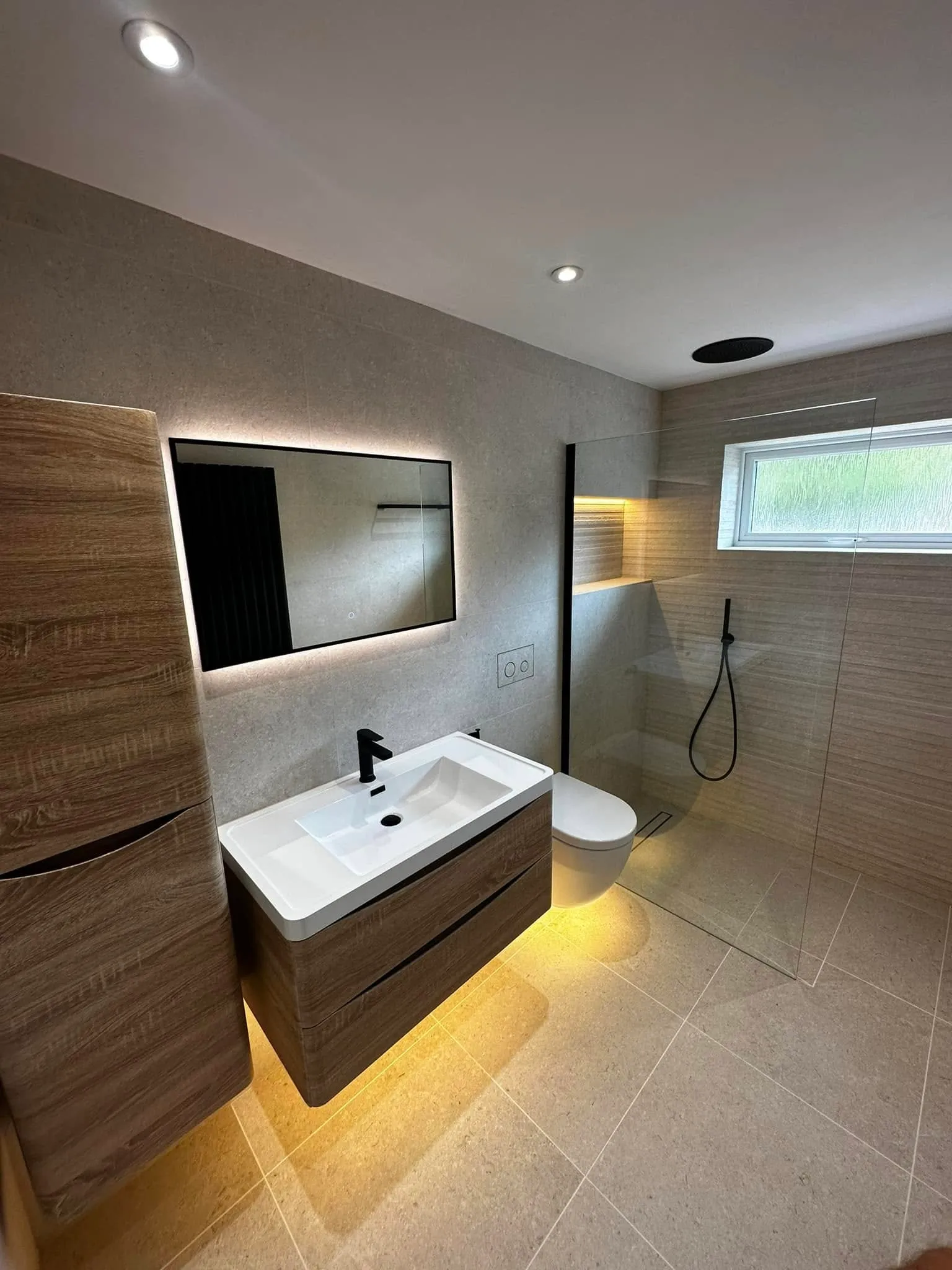 Wide view of the limestone en-suite: floating oak vanity, glowing mirror and walk-in shower