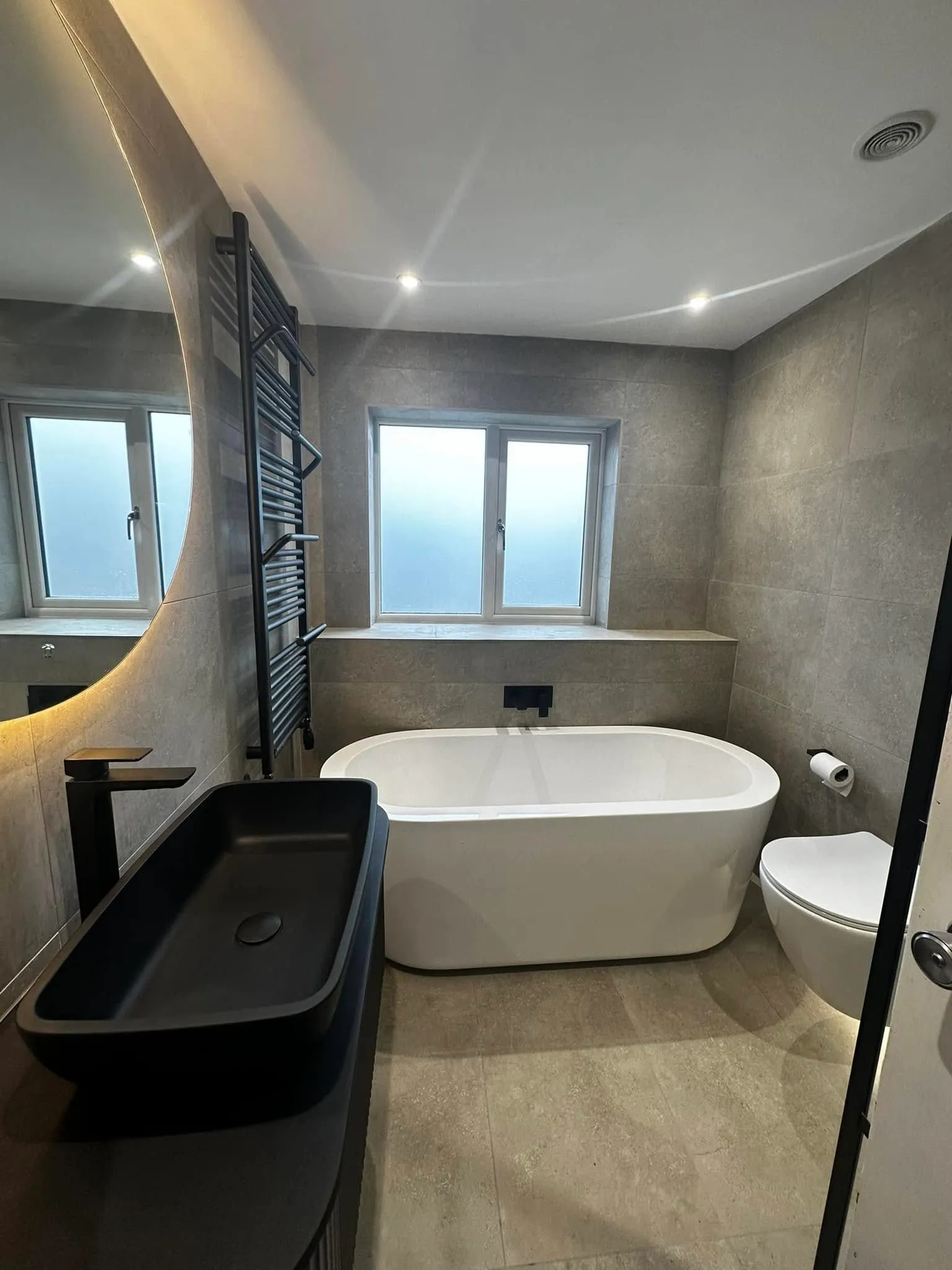 The finished bathroom — large-format grey stone porcelain, freestanding bath, matt black fittings and a backlit mirror